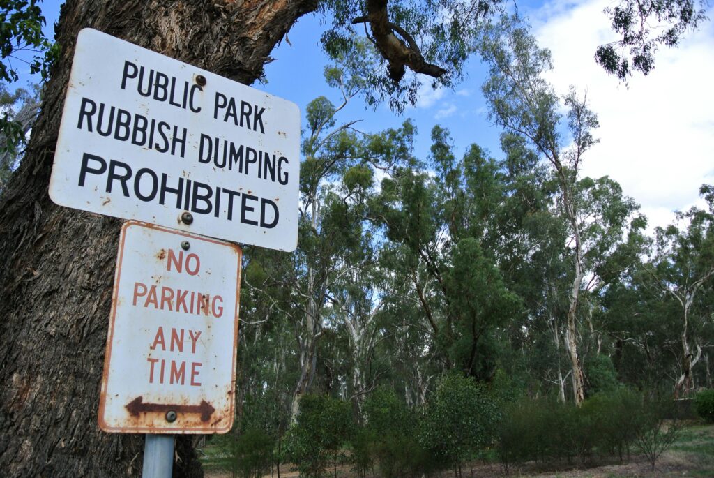 Rusty public park sign prohibiting rubbish dumping and parking at any time against a tree.