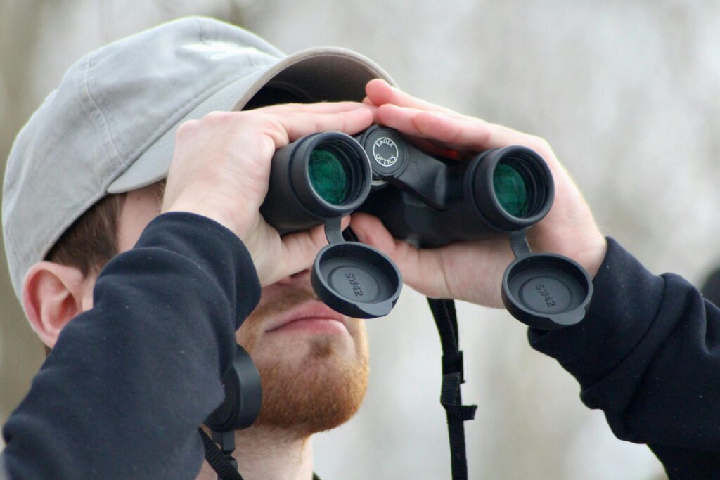 A young bearded man wearing a gray cap observing birds through binoculars in New Wilmington, Pennsylvania.